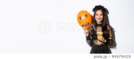 Image of cheerful asian girl trick or treating on halloween, holding sweets and orange balloon with scary face 94979429