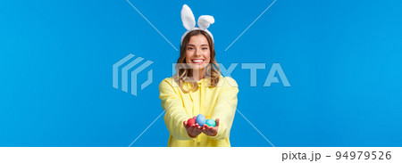 Holidays, people and fun concept. Charismatic cute blond girl holding coloured Easter eggs, wearing bunny ears and smiling joyfully, congratulate family with holy day, blue background Holidays, people and fun concept. Charismatic cute blond girl holding coloured Easter eggs, wearing bunny ears and smiling joyfully, congratulate family with holy day, blue background 94979526