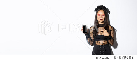 Young elegant woman in gothic dress and black wreath pointng finger at smartphone screen with pleased smile on her face, standing over white background Young elegant woman in gothic dress and black wreath pointng finger at smartphone screen with pleased smile on her face, standing over white background 94979688