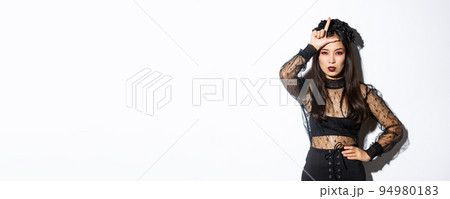 Image of arrogant sexy asian woman in witch costume showing loser gesture on forehead, mocking someone at halloween party, standing over white background 94980183