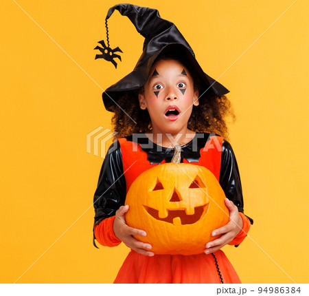 Happy cheerful ethnic girl in costume holding pumpkin while celebrates Halloween on yellow background Happy cheerful ethnic girl in costume holding pumpkin while celebrates Halloween on yellow background 94986384