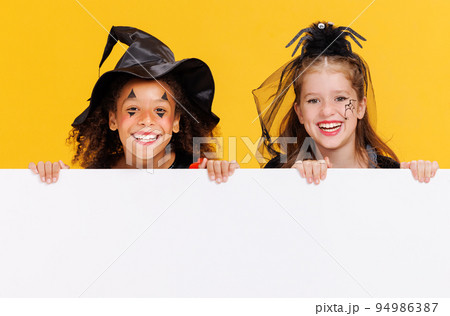 Happy Halloween. Kids girls in carnival costumes and makeup with blank white poster on colored yellow background 94986387