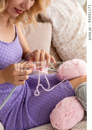 Close-up of woman in a lilac home dress knits a light pink scarf or plaid from natural threads on the bed. Close-up of woman in a lilac home dress knits a light pink scarf or plaid from natural threads on the bed. 94991171