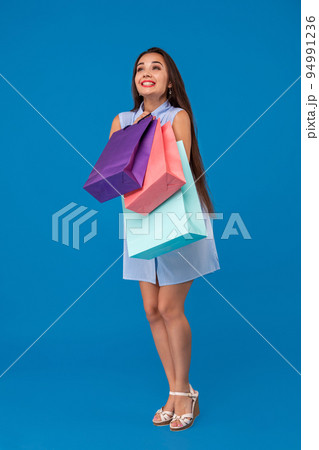 Beautiful young woman holding shopping bags and looking away while standing against blue background. 94991236