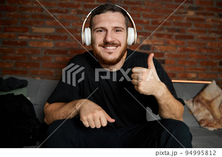 Handsome bearded man in headphones listening to music at home. Music. Half-length portrait of young guy in casual youth style clothes with sign ok sitting on sofa. Concept of emotions. Handsome bearded man in headphones listening to music at home. Music. Half-length portrait of young guy in casual youth style clothes with sign ok sitting on sofa. Concept of emotions. 94995812
