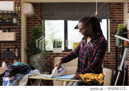 African american woman ironing clothes at home, laundry and ironing day, independent lifestyle in urban apartment, modern housewife doing chores and housework at home. Front view. 95006125