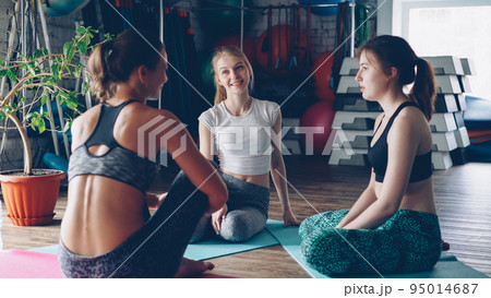 Young female yoga students are sitting on mats and talking while waiting for morning practice in large light gym. Fitness balls, step aerobic platforms and other sports equipment is visible. 95014687