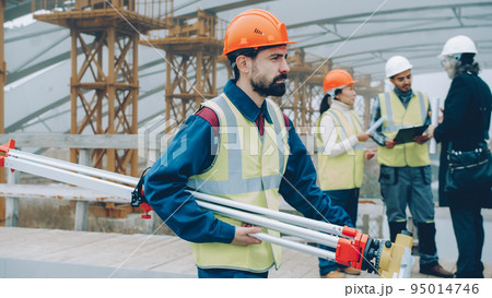 Side view of geodesy specialist walking in building area holding equipment wearing uniform on workday. Professionals and workplace concept. Side view of geodesy specialist walking in building area holding equipment wearing uniform on workday. Professionals and workplace concept. 95014746