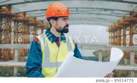 Portrait of serious foreman wearing uniform checking blueprint and looking at construction site outdoors in urban area. People and occupation concept. 95014773