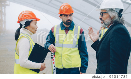 Builders wearing uniform are talking at construction site while architect in suit and helmet is coming discussing project gesturing and looking around Builders wearing uniform are talking at construction site while architect in suit and helmet is coming discussing project gesturing and looking around 95014785