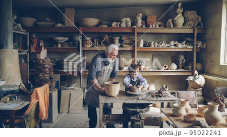 Experienced potter loving prandparent is teaching his young grandchild pottery in small home workshop. Boy is working with throwing wheel, grandpa is talking to him. Experienced potter loving prandparent is teaching his young grandchild pottery in small home workshop. Boy is working with throwing wheel, grandpa is talking to him. 95022241