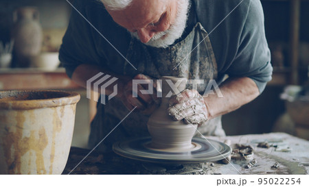 Close-up shot of half-finished ceramic jar spinning on potters's wheel and male hands molding clay with professional tools. Creating eathenware and traditional pottery concept. Close-up shot of half-finished ceramic jar spinning on potters's wheel and male hands molding clay with professional tools. Creating eathenware and traditional pottery concept. 95022254