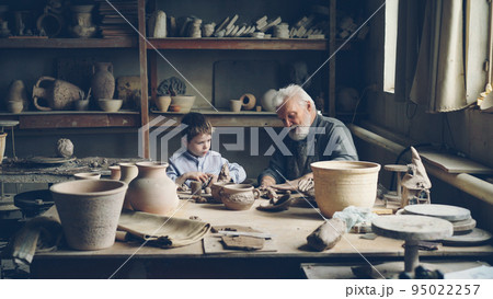 Skilled child is helping his senior grandfather professional potter to knead clay for handmade ceramic figures. Loving family, apprenticeship and conventional pottery concept. 95022257