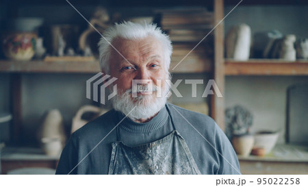 Close-up portrait of senior potter in muddy apron standing alone at table in workshop, smiling and looking at camera. Shelves with handmade vases and pots in background. 95022258