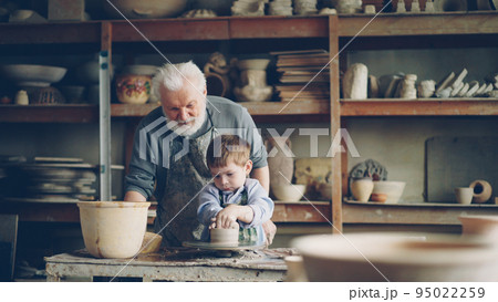 Caring grandfather experienced potter is teaching little boy how to work with clay on potter's wheel. Grandson is making mistake, loving patient grandpa is helping him. Caring grandfather experienced potter is teaching little boy how to work with clay on potter's wheel. Grandson is making mistake, loving patient grandpa is helping him. 95022259