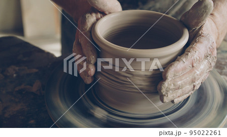 Senior male ceramist is concentrated on making pot from clay on spinning throwing wheel. He is correcting form of earthenware and finishing ornament. 95022261