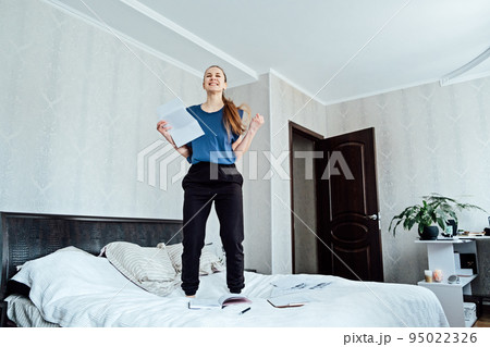 Excited cheerful young woman received a letter, she is hired, jumping on sofa at home, getting good news, feeling joy, dancing, laughing, making winner gesture, happy to win prize 95022326