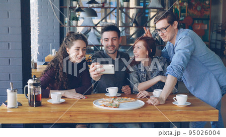 Young bearded man is making video call from pizza house together with best friends. Playful mates are talking, smiling, making funny faces and waving hands looking at camera. 95026004