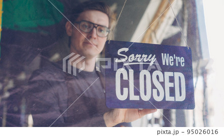 Handsome male cafe worker in apron is changing doorplate to sorry we are closed. End of work day, evening and catering business concept. Handsome male cafe worker in apron is changing doorplate to sorry we are closed. End of work day, evening and catering business concept. 95026016