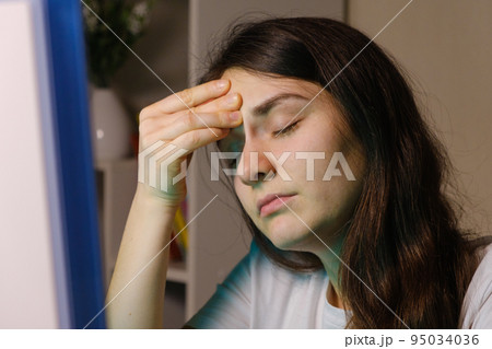 A tired woman holds her hand by the forehead, sitting in front of a computer screen A tired woman holds her hand by the forehead, sitting in front of a computer screen 95034036