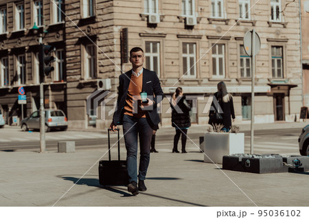 Going to airport terminal. Confident businessman traveler walking on city streets and pulling his suitcase drinking coffee and speaking on smartphone Going to airport terminal. Confident businessman traveler walking on city streets and pulling his suitcase drinking coffee and speaking on smartphone 95036102