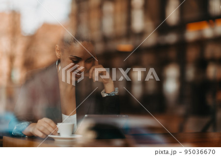 woman in a coffee shop drink coffee viewed through glass with reflections as they sit at a table chatting and laughing 95036670