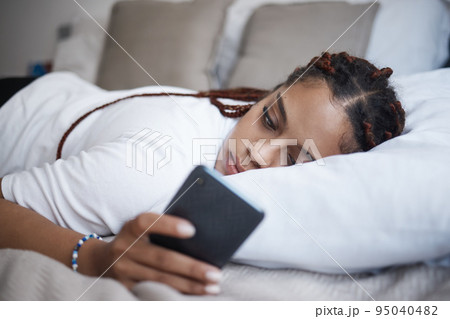 Depression, sad and african woman on a phone relaxing on the bed in her bedroom at home. Tired, mental health and lonely black girl browsing on social media or internet while having breakup problems. 95040482