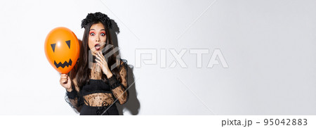 Portrait of surprised asian woman in halloween costume, dressed-up as witch, holding orange balloon with scary face Portrait of surprised asian woman in halloween costume, dressed-up as witch, holding orange balloon with scary face 95042883