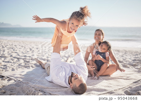 Family, children and playing with a girl on a beach holiday with her parents and sister during summer. Kids, travel and ocean with a female child or daughter on the sand by the sea with her father 95043668