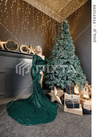 Portrait of woman posing for a photo. Gorgeous blonde girl in an elegant long green dress celebrates Christmas. Woman standing by a beautiful Christmas tree in expensive interiors. 95045099