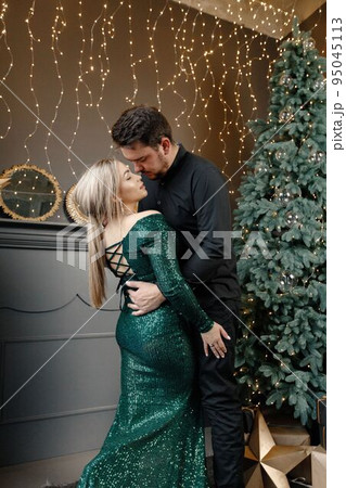 Young woman and man standing near christmas tree and kissing. Cheerful family celebrating Christmas. Blonde woman wearing fashion green dress and bearded man wearing black shirt. 95045113
