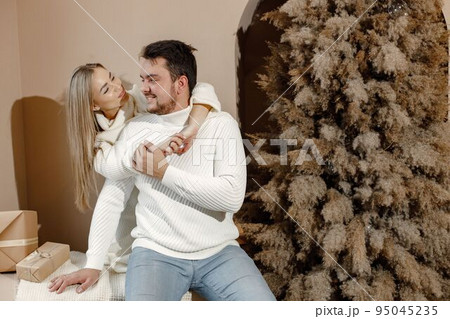 Young woman and man sitting near christmas tree and hugging. Cheerful family celebrating Christmas. Blonde woman and bearded man wearing white clothes. 95045235
