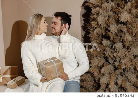 Young woman and man sitting near christmas tree with a gift box. Cheerful family celebrating Christmas. Blonde woman and bearded man wearing white clothes. 95045242
