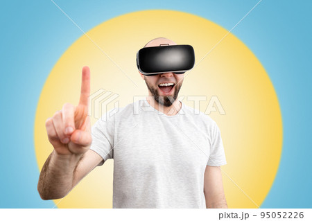 VR glasses. A wondered man in virtual reality glasses, with his mouth slightly open, points a finger into space. Blue background with a yellow circle. The concept of virtual reality VR glasses. A wondered man in virtual reality glasses, with his mouth slightly open, points a finger into space. Blue background with a yellow circle. The concept of virtual reality 95052226