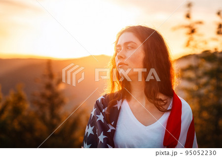Independence day and Memorial Day. Portrait of Caucasian young woman with American flag at shoulders. Sunset in the background. The concept of American National Holidays 95052230