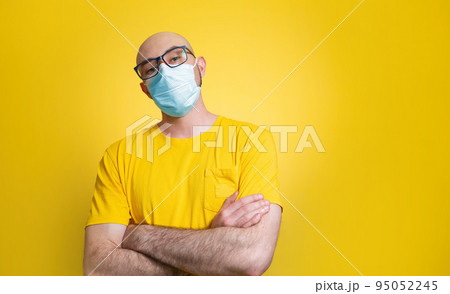 Against a yellow background stands a bald man in a yellow t-shirt, glasses and a medical mask, confidently crossing his arms over his chest. Concept of quarantine, self-isolation and coronavirus 95052245