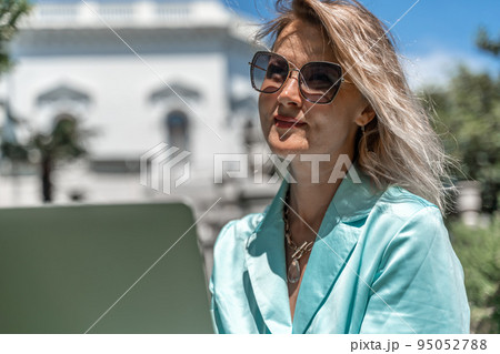 a young beautiful woman with blond curly hair in glasses and a blue dress sits on the grass in nature and uses a laptop, 95052788