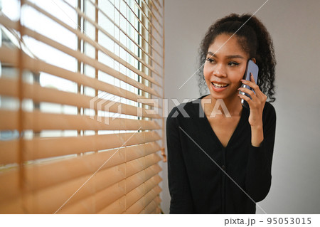 Smiling young woman standing near window in home office and talking on mobile phone Smiling young woman standing near window in home office and talking on mobile phone 95053015