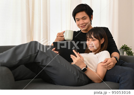 Loving asian couple embracing and browsing internet on digital tablet in bright living room. People and technology concept Loving asian couple embracing and browsing internet on digital tablet in bright living room. People and technology concept 95053016