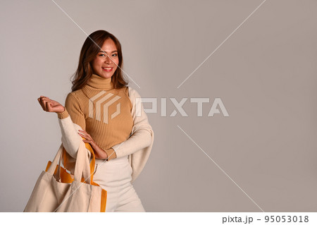 Portrait of attractive young woman wearing warm clothes holding white tote bag standing over grey background Portrait of attractive young woman wearing warm clothes holding white tote bag standing over grey background 95053018