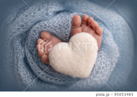 The tiny foot of a newborn baby. Soft feet of a new born in a blue wool blanket. Close up of toes, heels and feet of a newborn. Knitted white heart in the legs of a baby. Macro photography. The tiny foot of a newborn baby. Soft feet of a new born in a blue wool blanket. Close up of toes, heels and feet of a newborn. Knitted white heart in the legs of a baby. Macro photography. 95053465