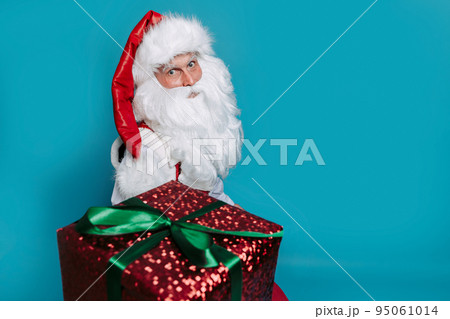 Santa Claus gives xmas gift box while looking at camera on blue studio background. New Year concept 95061014