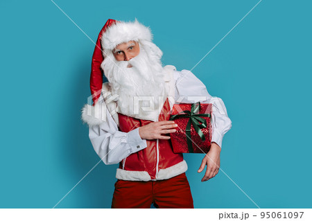 Christmas. Stylish santa claus holds red gift box on color background while looking at camera. 95061097