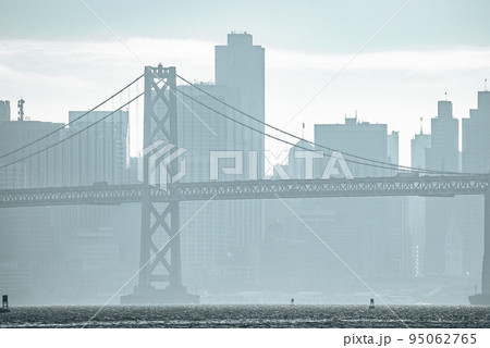 Idyllic view of Bay Bridge and urban skyline during foggy weather in San Francisco. Downtown district lying on sea front with cloudy sky in background. Famous tourist attraction in city. 95062765