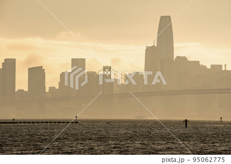 Bay Bridge and downtown district on coastal seafront. Famous tourist attraction connecting urban San Francisco. Scenic view of city skyline with sky in background during sunset. 95062775