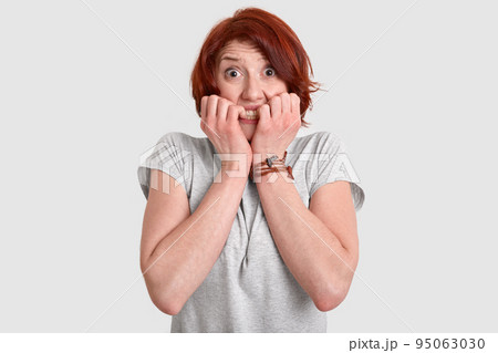 Photo of frightened red haired woman bites fingers nails, looks with nervous expression, afraids of difficulties, dressed in casual t shirt, models over white background. Negative feelings concept Photo of frightened red haired woman bites fingers nails, looks with nervous expression, afraids of difficulties, dressed in casual t shirt, models over white background. Negative feelings concept 95063030