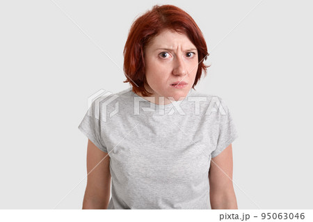 Photo of beautiful redhead woman looks with curious gaze, bewilderment at camera, wears casual t shirt, models over white studio background, has displeased facial expression. People and emotions 95063046