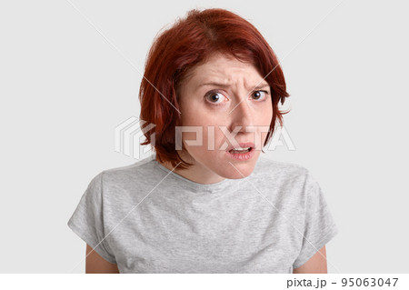 Close up shot of pretty young woman has short foxy hair, looks scrupulously with frustrated gaze, dressed in casual clothes, being puzzled by hearing something negative, models over white studio wall 95063047