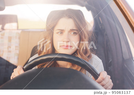 Nervous female driver sits at wheel, has worried expression as afraids to drive car by herself for first time. Frightened woman has car accident on road. People, driving, problems with transport 95063137