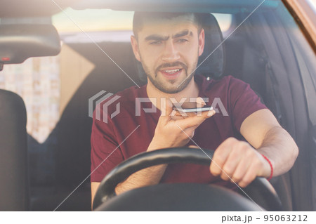 Bearded young male with thick stubble makes voice call via cell phone, drives car on high speed, risks as has distracted attention, sits in front of wheel. People, technology and driving concept 95063212